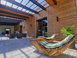 Uma área externa coberta com piso de madeira, um banco de madeira com almofada xadrez e um vaso de planta.