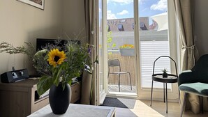 Ein Zimmer mit einer Schiebeglastür, die auf einen Balkon führt, einem schwarzen Radio auf einem Schreibtisch, einem grünen Stuhl und einer Vase mit Sonnenblumen.