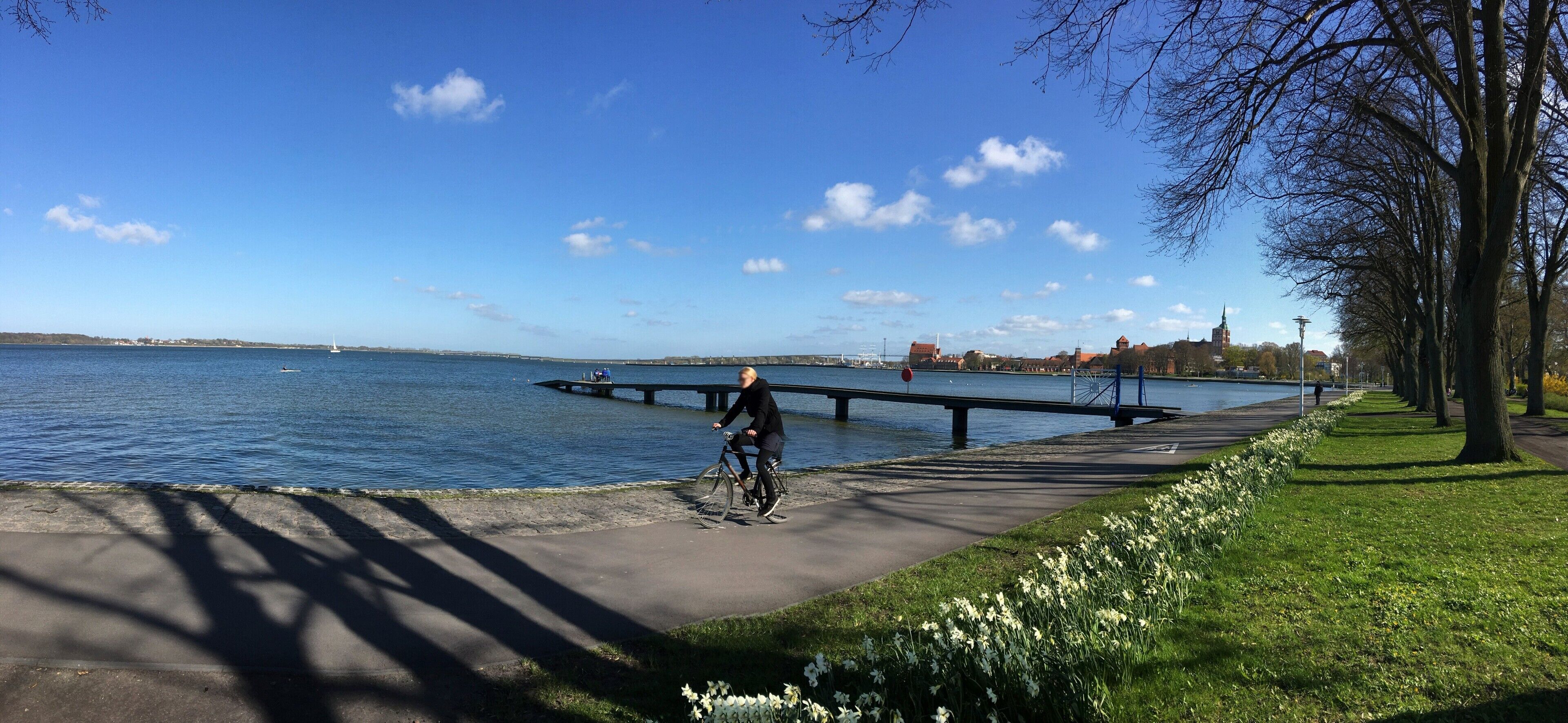 Une promenade pavée le long d’un plan d’eau, avec un cycliste, un pont et un bâtiment situé au loin.