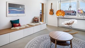 A modern living room with a wooden bench, a round table, a rocking chair, a guitar, and a hanging lamp.