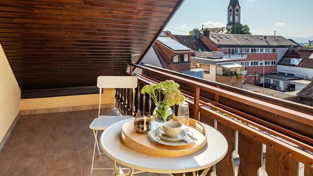 A balcony with a table, chairs, a vase with flowers, a tray with a cup and saucer, and a view of buildings and a church steeple.
