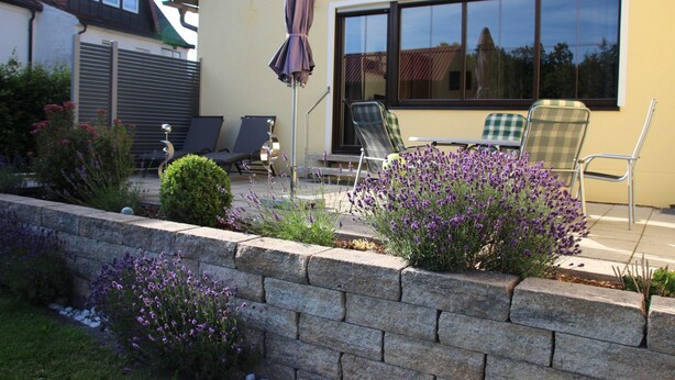 Un patio con una mesa y sillas, un muro de contención de piedra y flores moradas.