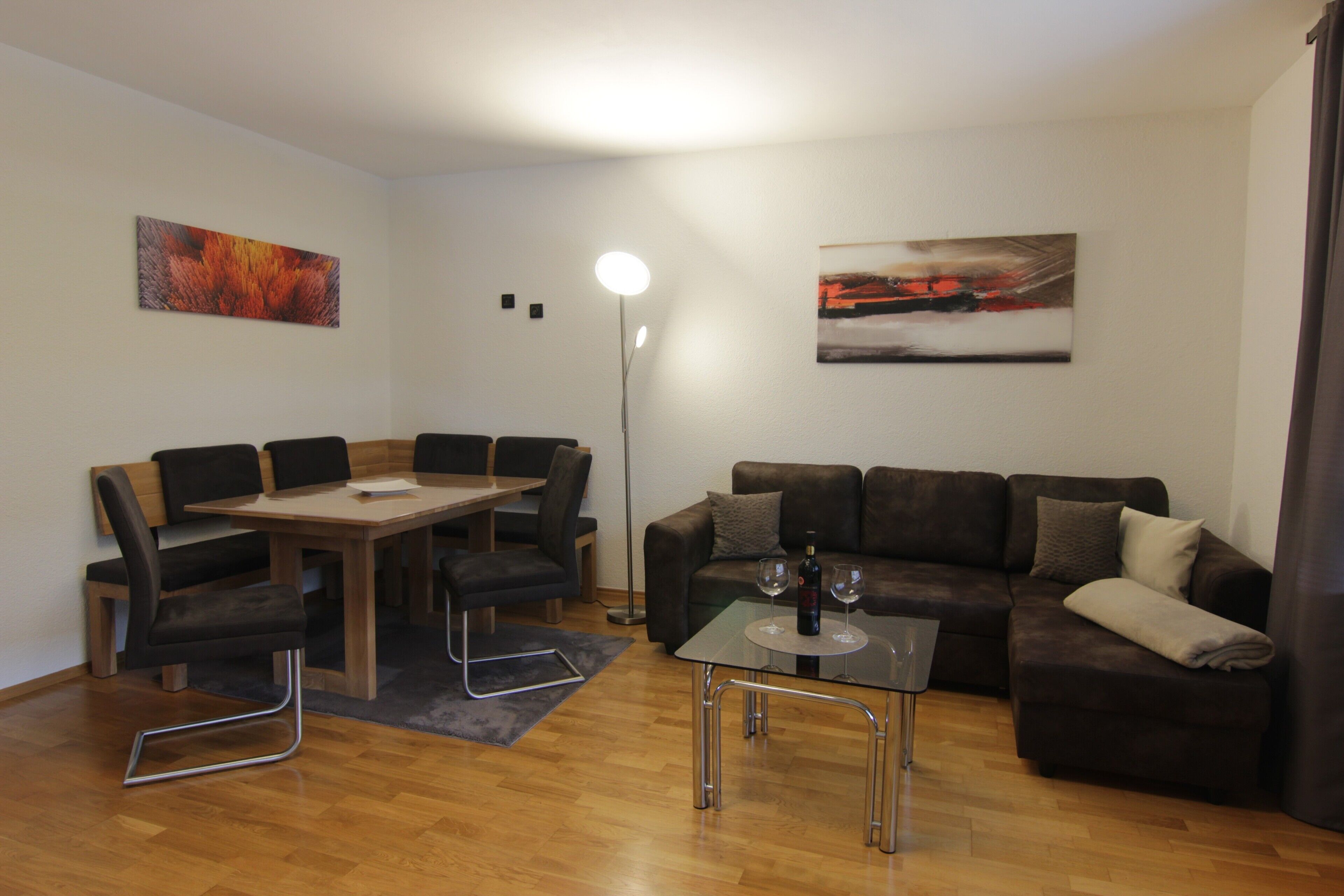 Een moderne woonkamer met een eettafel, stoelen, een glazen salontafel, een bank en twee abstracte schilderijen aan de wanden.