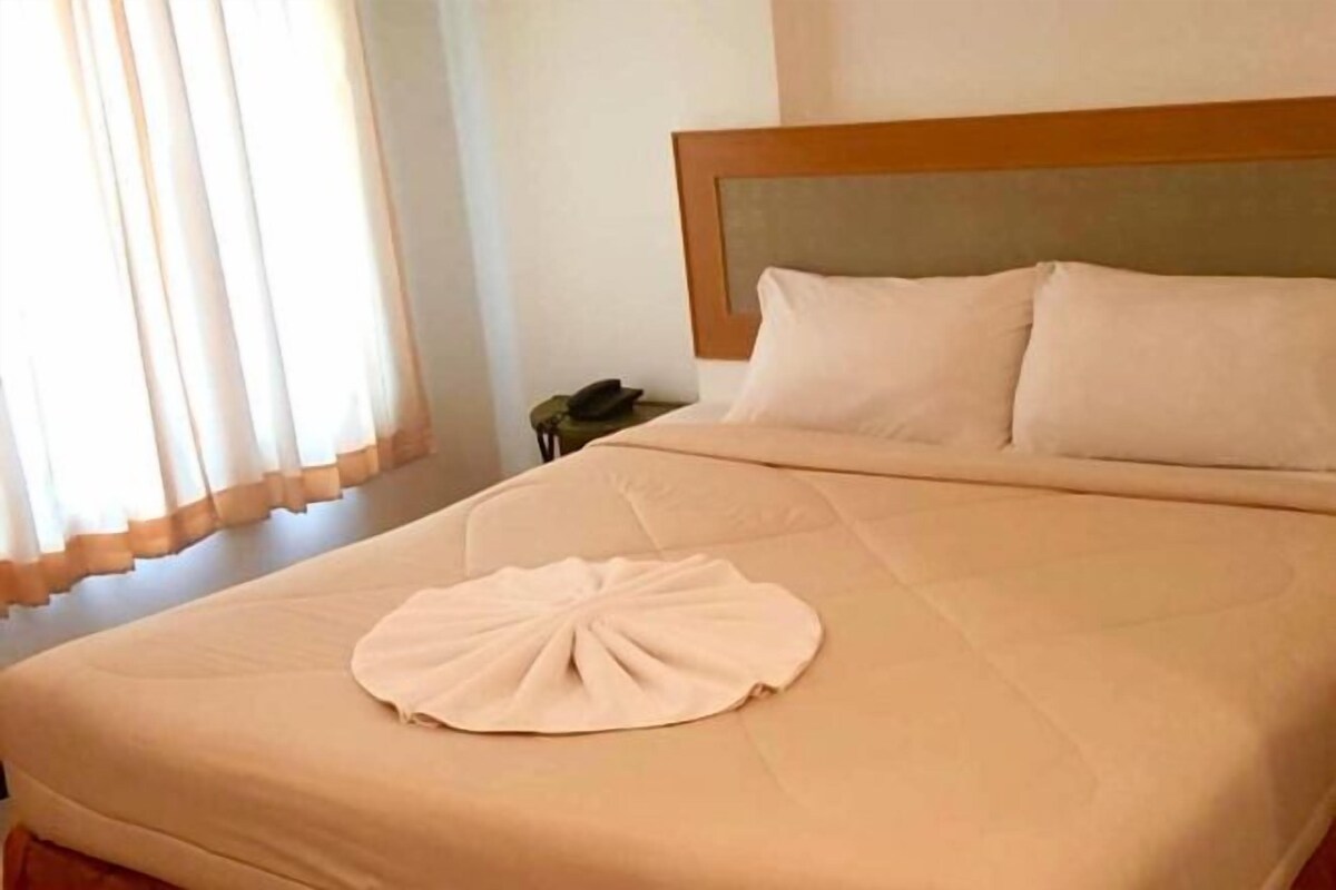 A neatly made bed with a headboard, a bedside table with a phone, and a window with curtains.