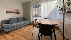 Ein modernes Wohnzimmer mit einem grauen Sofa, einem runden Holztisch mit Stühlen, einer Stehlampe und einem großen Fenster mit Blick auf Gebäude.