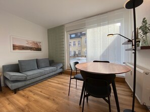 A modern living room with a grey sofa, a round wooden table with chairs, a floor lamp, and a large window with a view of buildings outside.