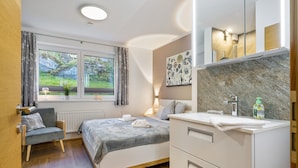 Une chambre à coucher comprenant un lit, une chaise, une table de chevet, un miroir, une fenêtre avec des rideaux et une vue sur la verdure à l’extérieur.