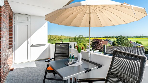 Un balcón con mesa, sillas, una sombrilla color beige y vistas a un paisaje verde.