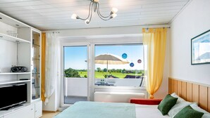 Habitación de hotel con cama, televisor, balcón con vistas a zonas verdes y ventana con cortina.