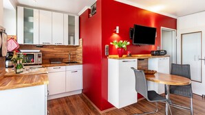 Cocina compacta con armarios blancos, una pared de acento roja, microondas, lavamanos, un pequeño comedor con mesa y sillas, y un televisor de pantalla plana montado sobre la encimera.