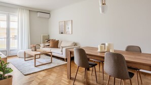 A modern living room with a wooden dining table, grey chairs, a white sofa, and a large window with curtains. - Graceful 2BR Apartment in Paleo Faliro (Paleo Faliro)