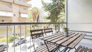 A balcony with wooden benches and a potted plant, overlooking a street with parked cars and buildings. - Graceful 2BR Apartment in Paleo Faliro (Paleo Faliro)