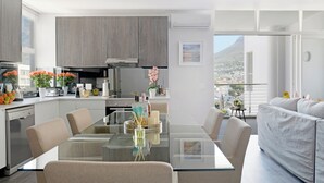 Una cuina moderna amb armaris grisos, una taula de dinar de vidre i un sofà gris en una zona d'estar àmplia i ben il·luminada amb vista a la ciutat.