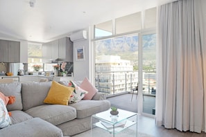 Un salon moderne avec un canapé gris, une table basse en verre et une vue sur un balcon donnant sur un paysage urbain et des montagnes en arrière-plan.