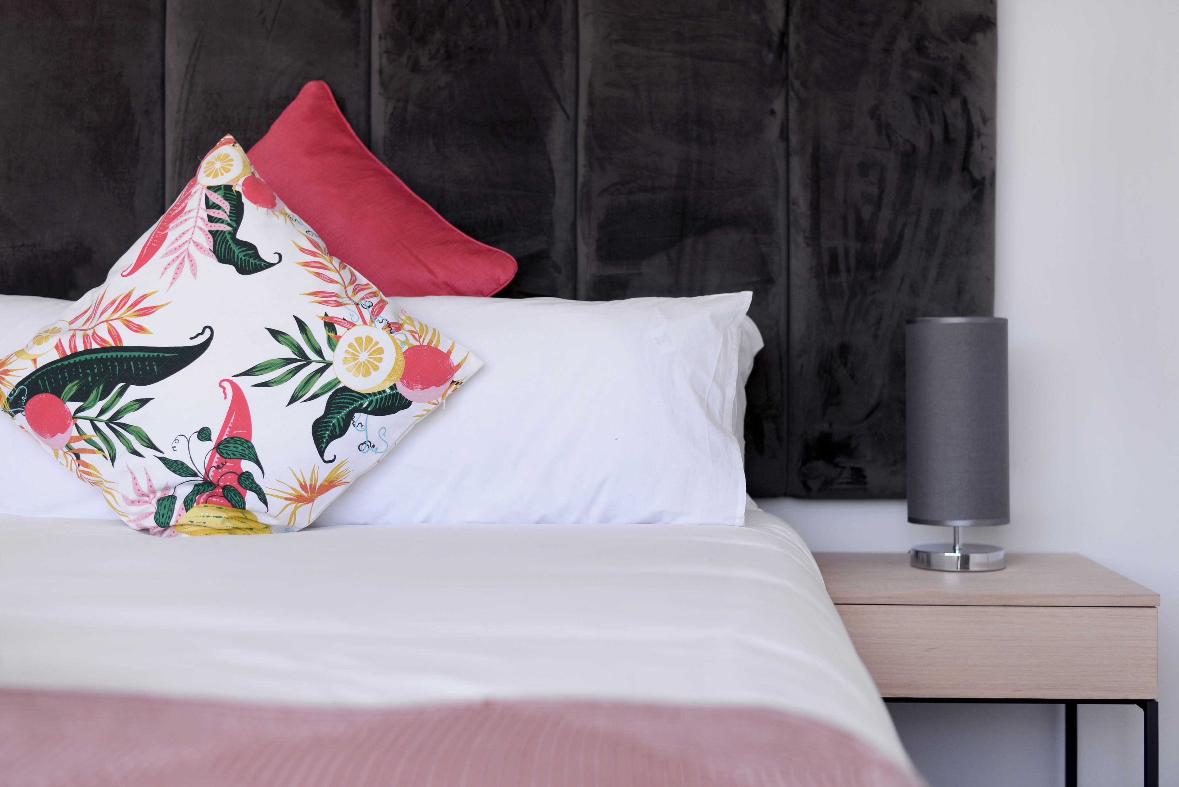 Una cama bien tendida con ropa de cama blanca y rosa, un cojín decorativo con estampado tropical y una mesita de noche con una lámpara.