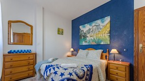 A bedroom with a bed, two bedside tables, a wooden dresser, a mirror, and a painting of a mountain lake. - Sea View Apartment Blue Pearl - Vista del Guanche (Arico)