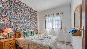 A bedroom with a floral wallpaper, a bed with a patterned bedspread, a bedside table with a lamp, a standing fan, and a wooden nightstand.