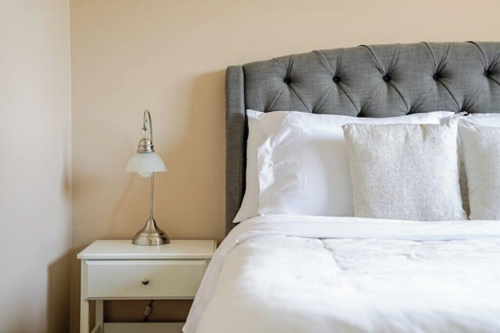 A neatly made bed with a grey upholstered headboard, a bedside lamp, and a white nightstand.
