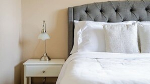 A neatly made bed with a grey upholstered headboard, a bedside lamp, and a white nightstand. - Tranquil Home in Bracewood Community of Calgary Southwest Alberta. (Calgary)