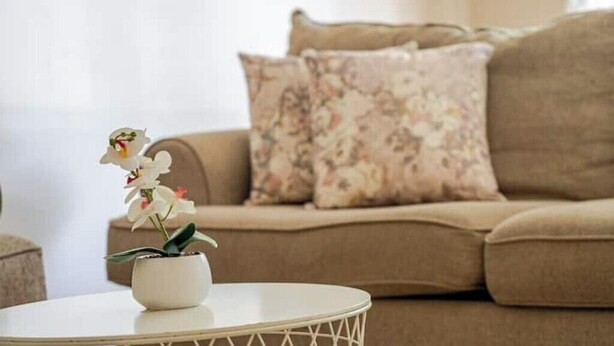 A beige sofa with floral cushions, a white round table with a potted orchid, and a white wall. - Tranquil Home in Bracewood Community of Calgary Southwest Alberta. (Calgary)