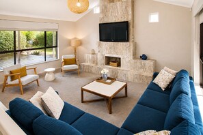 A living room with a blue sofa, a wooden coffee table, a white armchair, a lamp, and a TV mounted on a stone wall. - Gifford Getaway (Dunsborough)