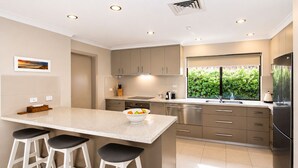 A modern kitchen with a central island, bar stools, stainless steel appliances, and a view of greenery through a window. - Gifford Getaway (Dunsborough)