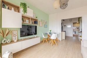Un salon moderne doté d’un téléviseur à écran plat, d’une table à manger avec des chaises, d’une cuisine en arrière-plan et d’un couloir menant à une autre pièce.
