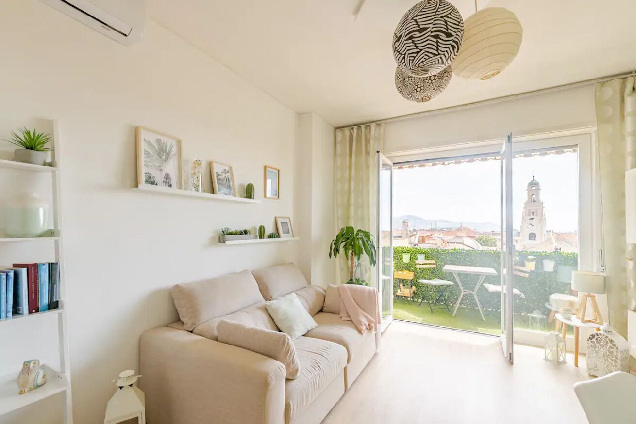 Una sala de estar moderna con un sofá, una estantería, una planta en maceta y vistas a un paisaje urbano a través de amplios ventanales.