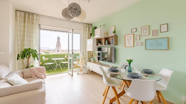 A modern living room with a dining area, a large window with a view of a tower, and a green wall with framed pictures.