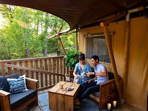 A wooden deck with a table and chairs, a covered area with a view of trees, and a window with curtains.