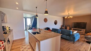 Et moderne køkken med en central ø, et spiseområde med en blå sofa og en stue med tv og pejs.