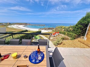 Une terrasse avec une table dressée pour prendre un verre, donnant sur une plage où l’on aperçoit des bateaux et des parasols.