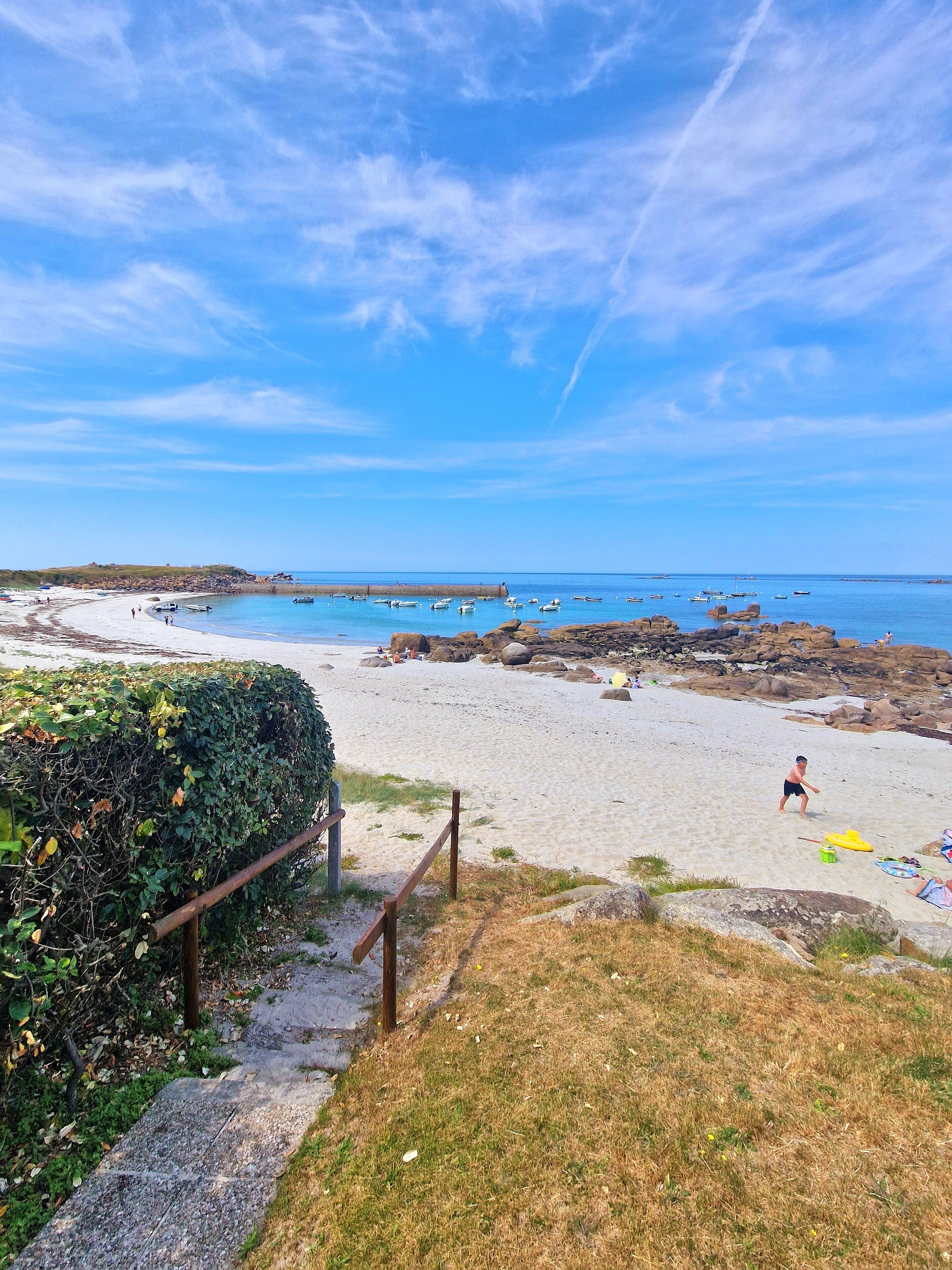 Una playa con cielo despejado, costa rocosa y barcos en el agua.