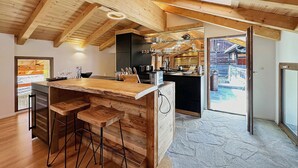Une cuisine moderne avec des poutres en bois, un coin bar avec des tabourets et une vue sur un paysage enneigé par la porte ouverte.