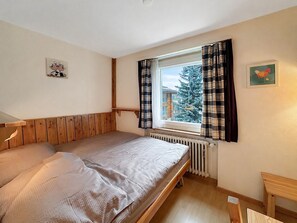 Una cama individual con cabecera de madera, una ventana con cortinas escocesas, un radiador y un cuadro pequeño en la pared.