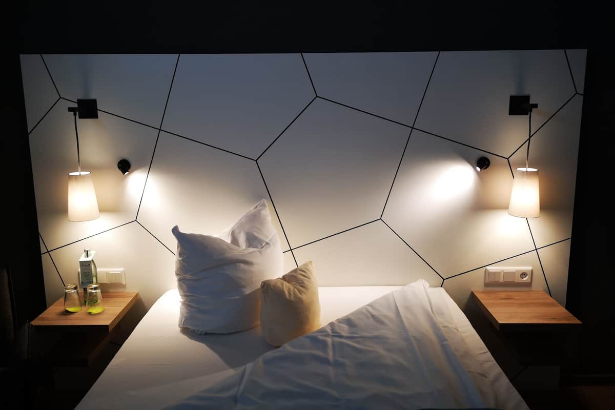 A modern hotel room with a geometric-patterned headboard, two bedside lamps, a wooden nightstand, and a bottle of water.