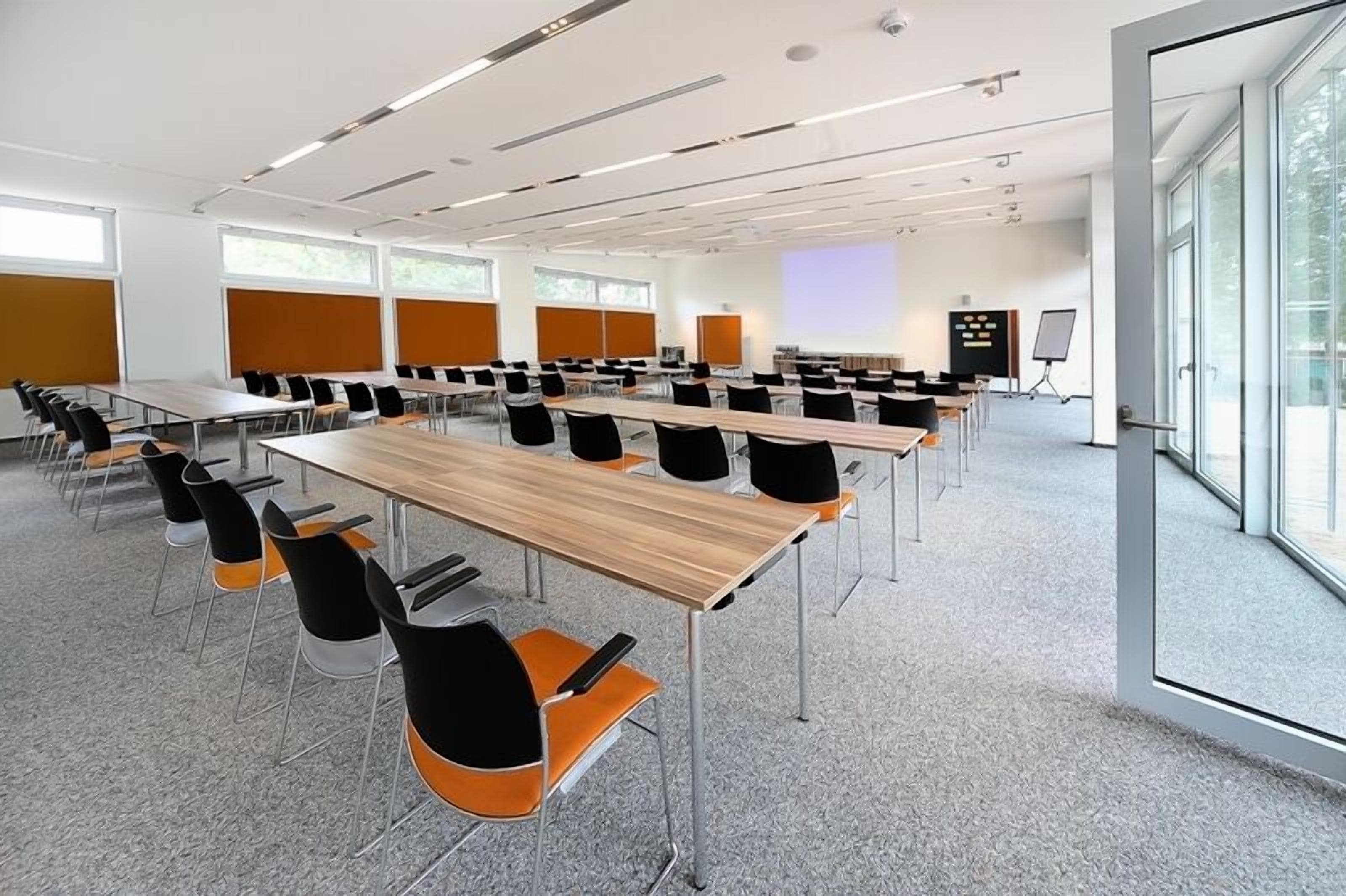 Un salón de conferencias con varias mesas y sillas, ventanas amplias y una pantalla de proyección.