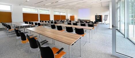 Un salón de conferencias con varias mesas y sillas, ventanas amplias y una pantalla de proyección.