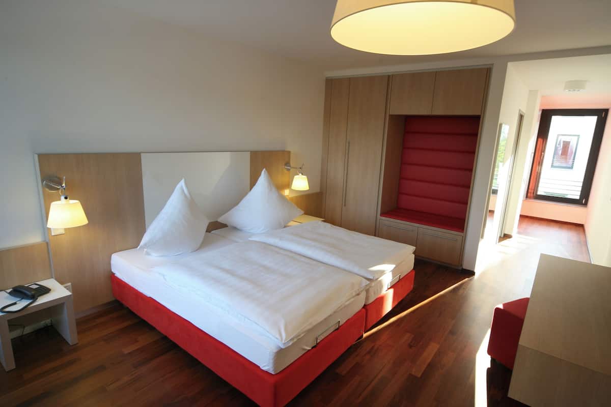 A modern hotel room with a large bed, two bedside lamps, a wooden headboard, and a red accent wall.
