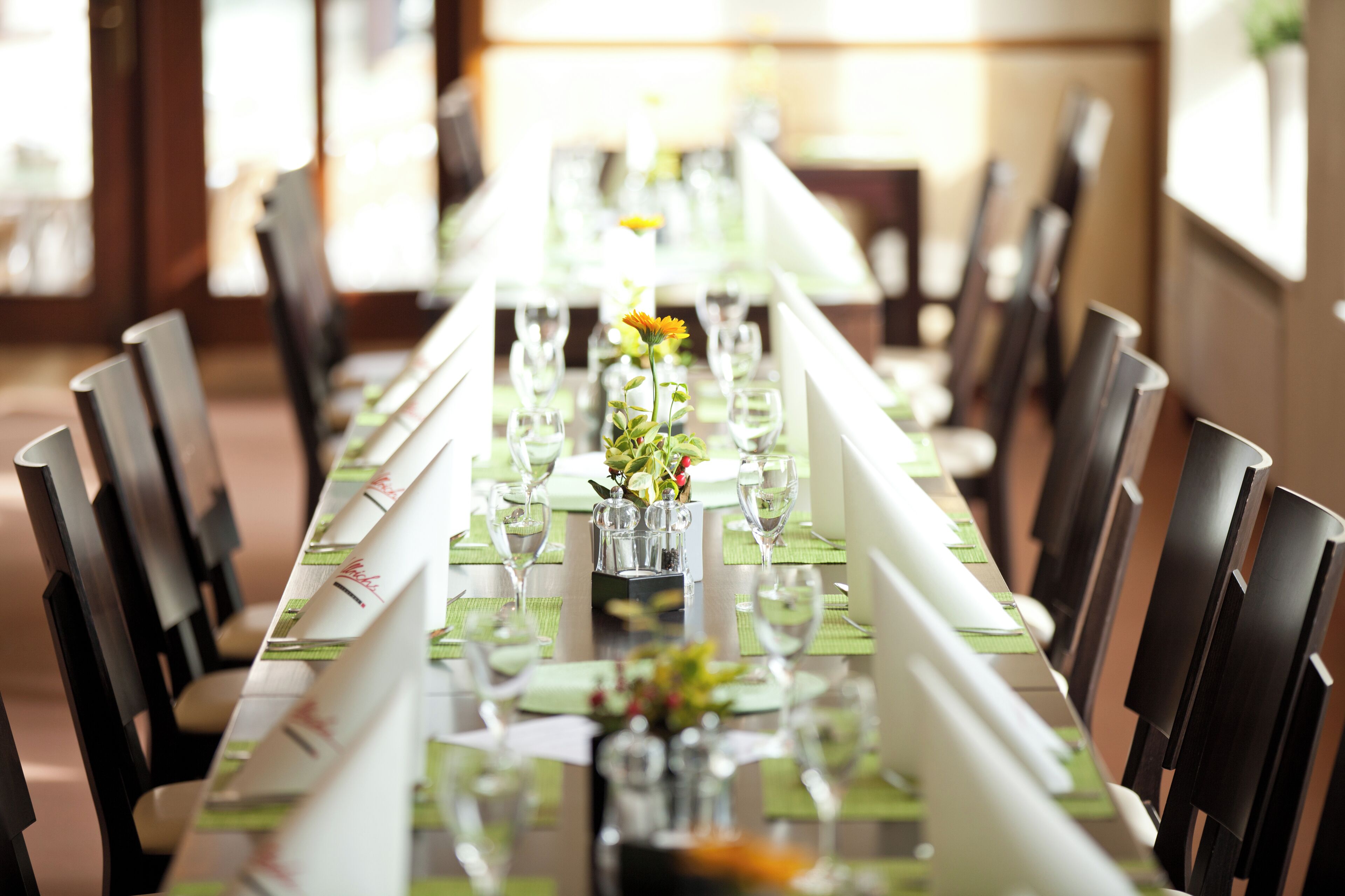 Una larga mesa de comedor dispuesta con sillas, caminos de mesa verdes y cubertería de vidrio transparente.