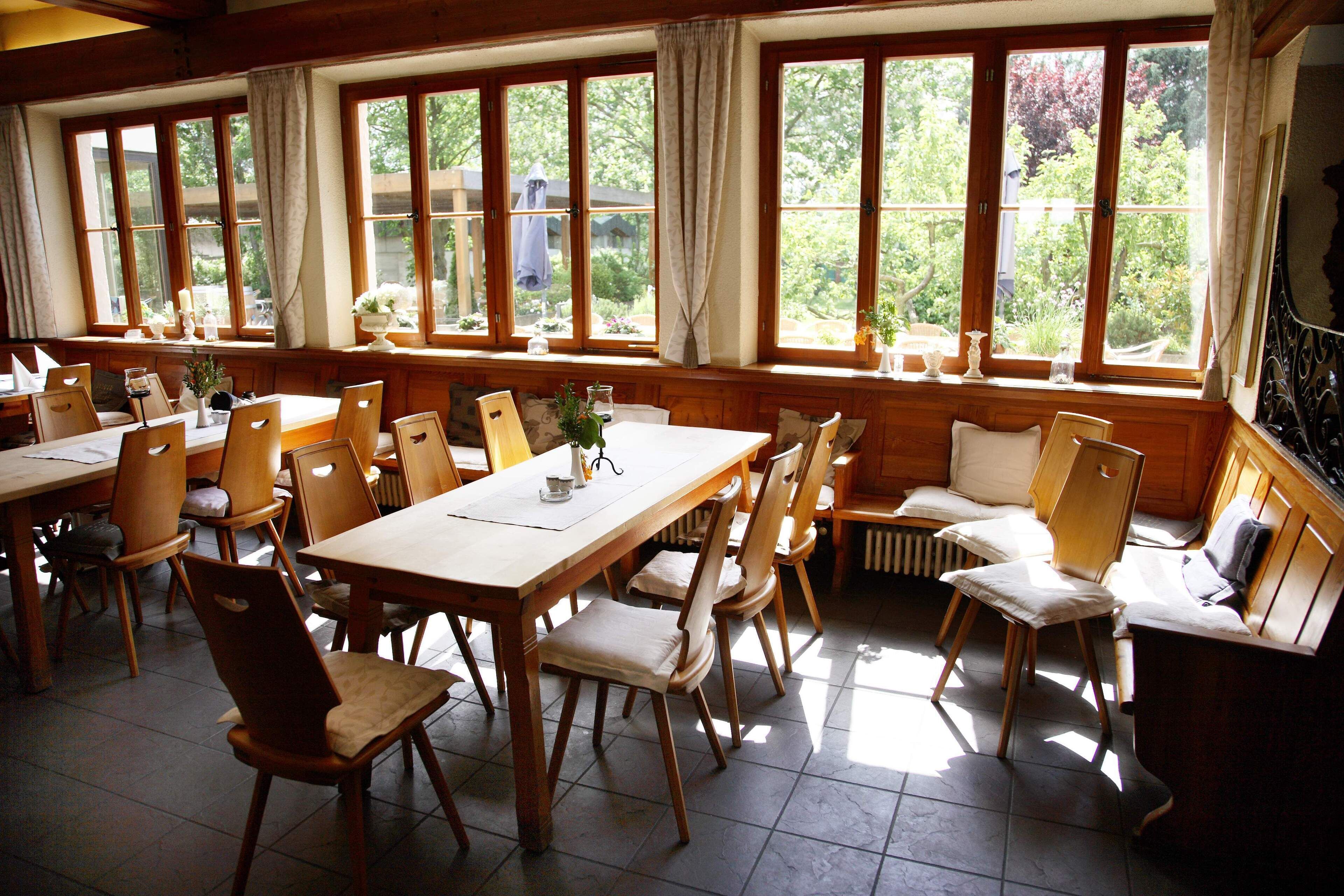 A dining area with wooden tables and chairs, large windows with curtains, and a view of greenery outside.