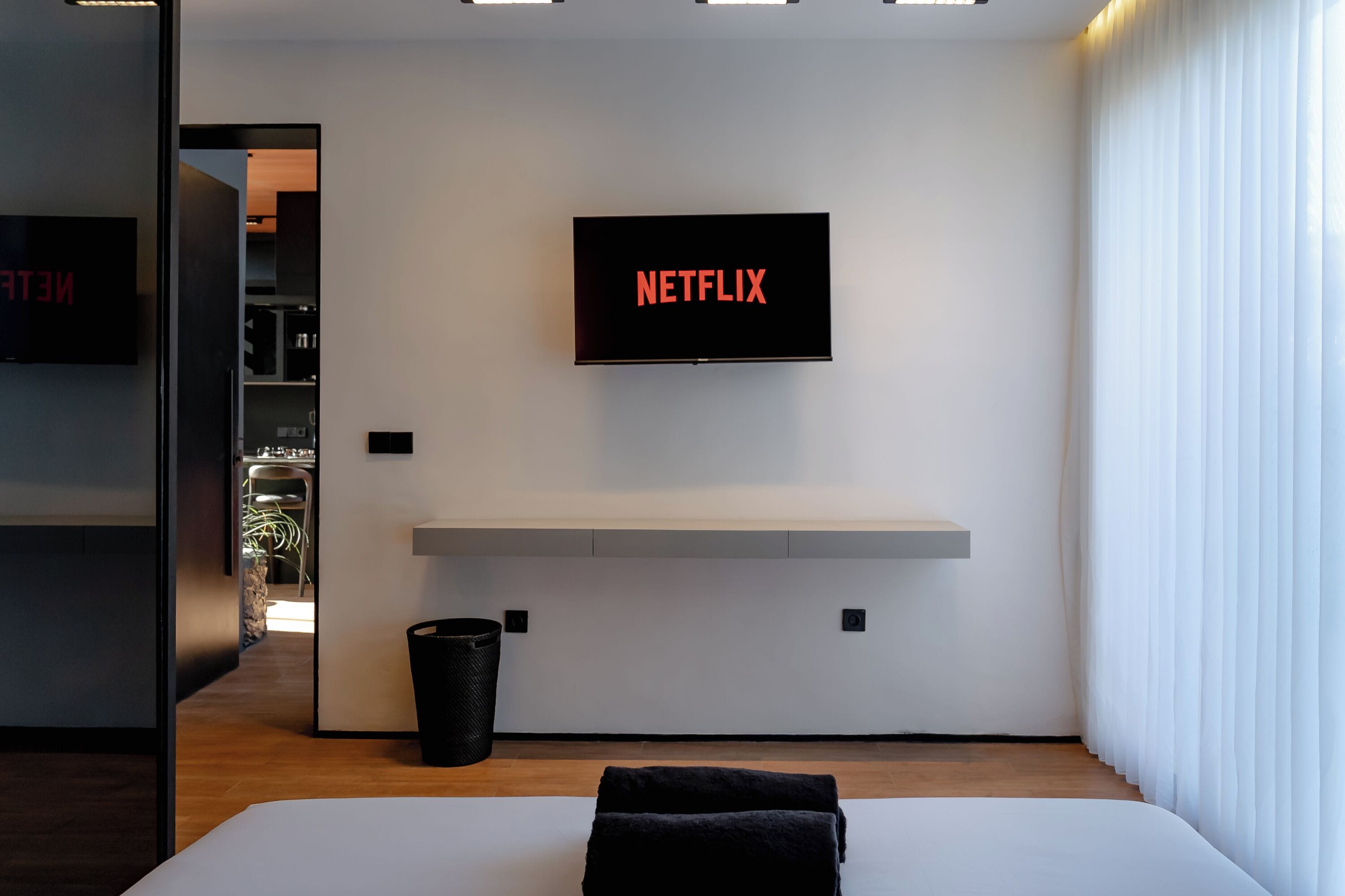 A modern hotel room with a large bed, a mounted TV displaying 'NETFLIX', a small shelf, and a waste bin.