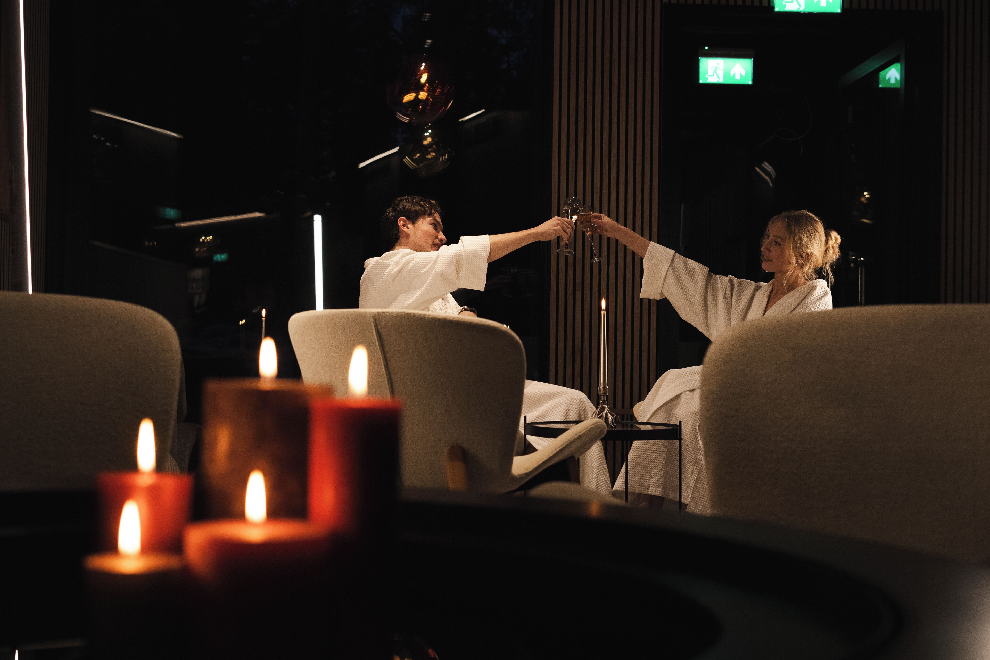 A dimly lit room with two people toasting, a table with candles, and a modern interior design.