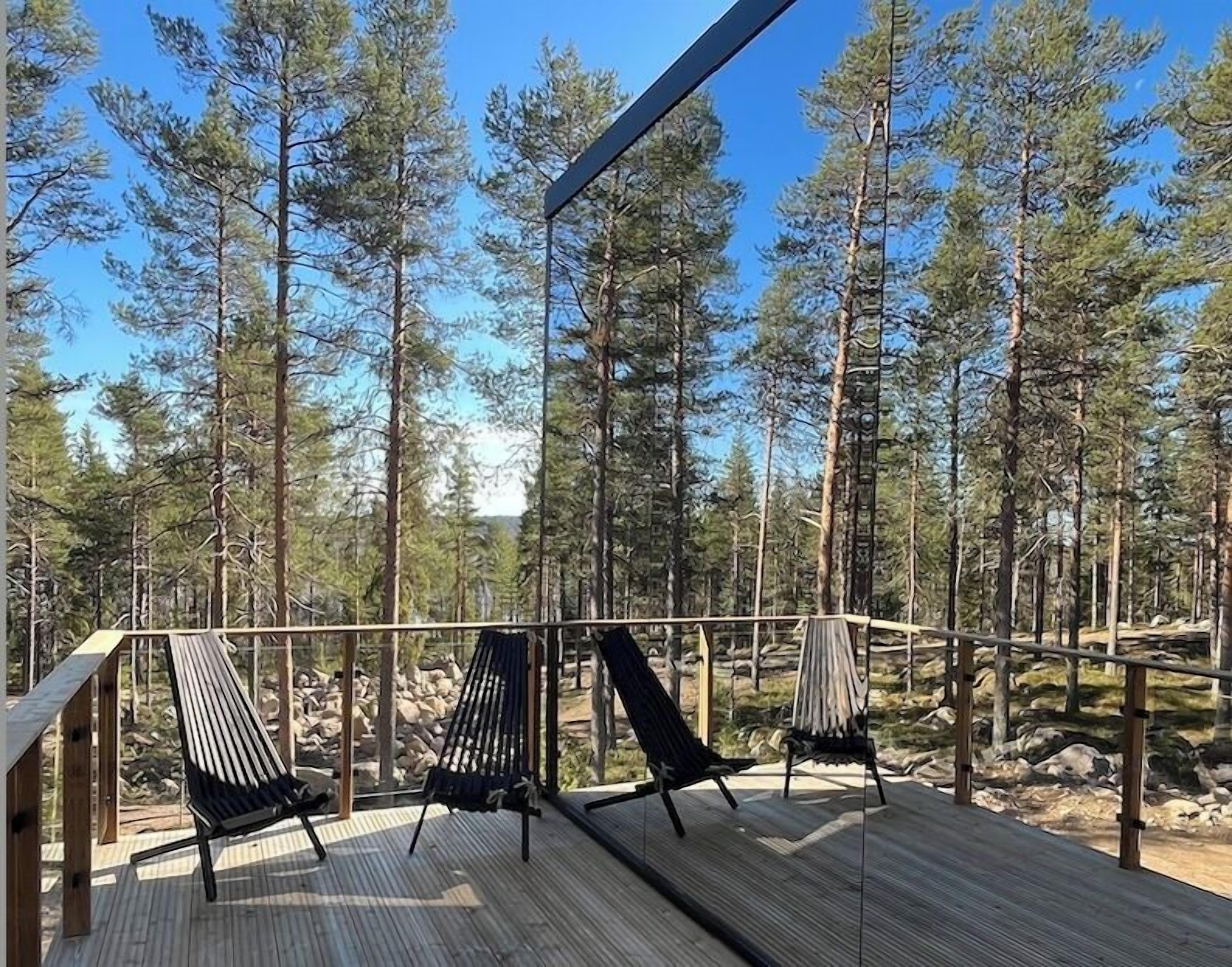 Puinen terassi, jossa on kaksi mustaa riippukeinua ja tuoli, ja sen ympärillä kohoaa korkeita mäntyjä.