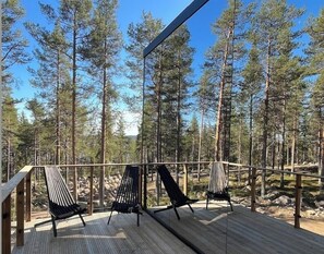 A wooden deck with two black hammocks and a chair, surrounded by tall pine trees. - Invisible Forest Lodge (Rovaniemi)