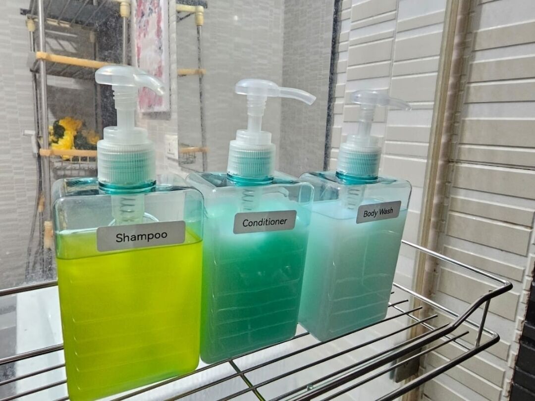 Tres botellas de líquido sobre una estantería con las etiquetas 'Champú', 'Acondicionador' y 'Gel de ducha'.