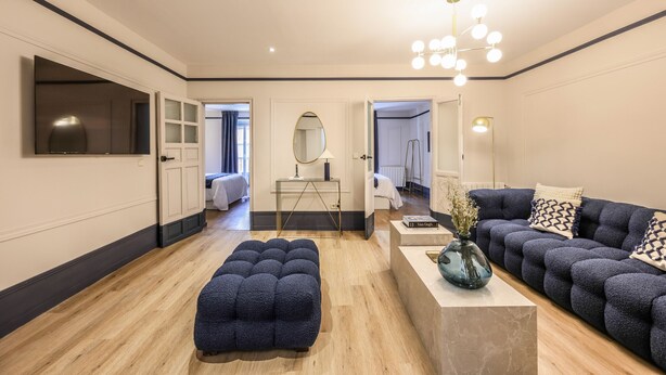 A modern living room with a sofa, ottoman, and a coffee table. There is a mirror, a lamp, and a TV mounted on the wall.