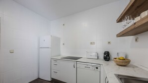 A compact kitchen with white cabinets, a dishwasher, a microwave, and a wooden shelf with glasses.