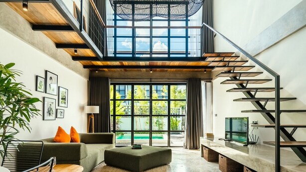 A spacious living area with a high ceiling, a staircase, and large windows offering a view of the outdoors. - Berawa Loft 10 by Betterplace (Canggu)