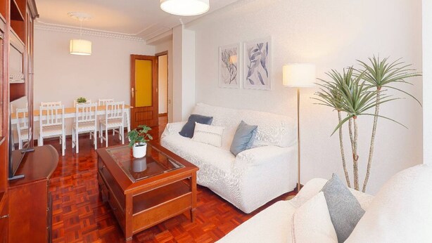 A living room with a white sofa, wooden coffee table, and a dining area with white chairs. - 1A130 Coqueto apartamento cerca playa (Gijón)